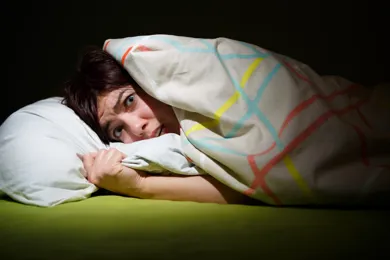 En kvinde ligger i sin seng og gemmer sig under sin dyne, fordi hun er nervøs og bange.